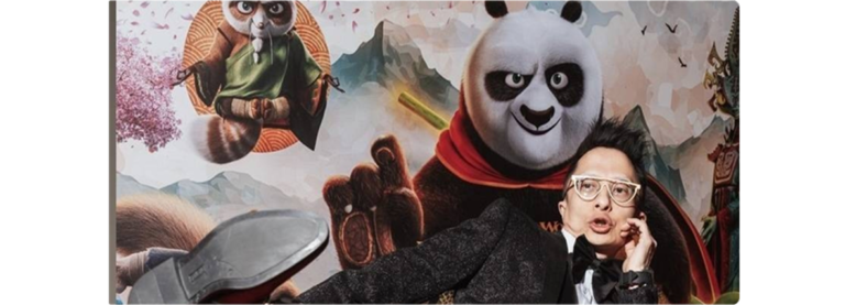 Kwok One in a black tuxedo with white glasses poses playfully in front of a "Kung Fu Panda" poster, featuring the character Po in a martial arts stance.